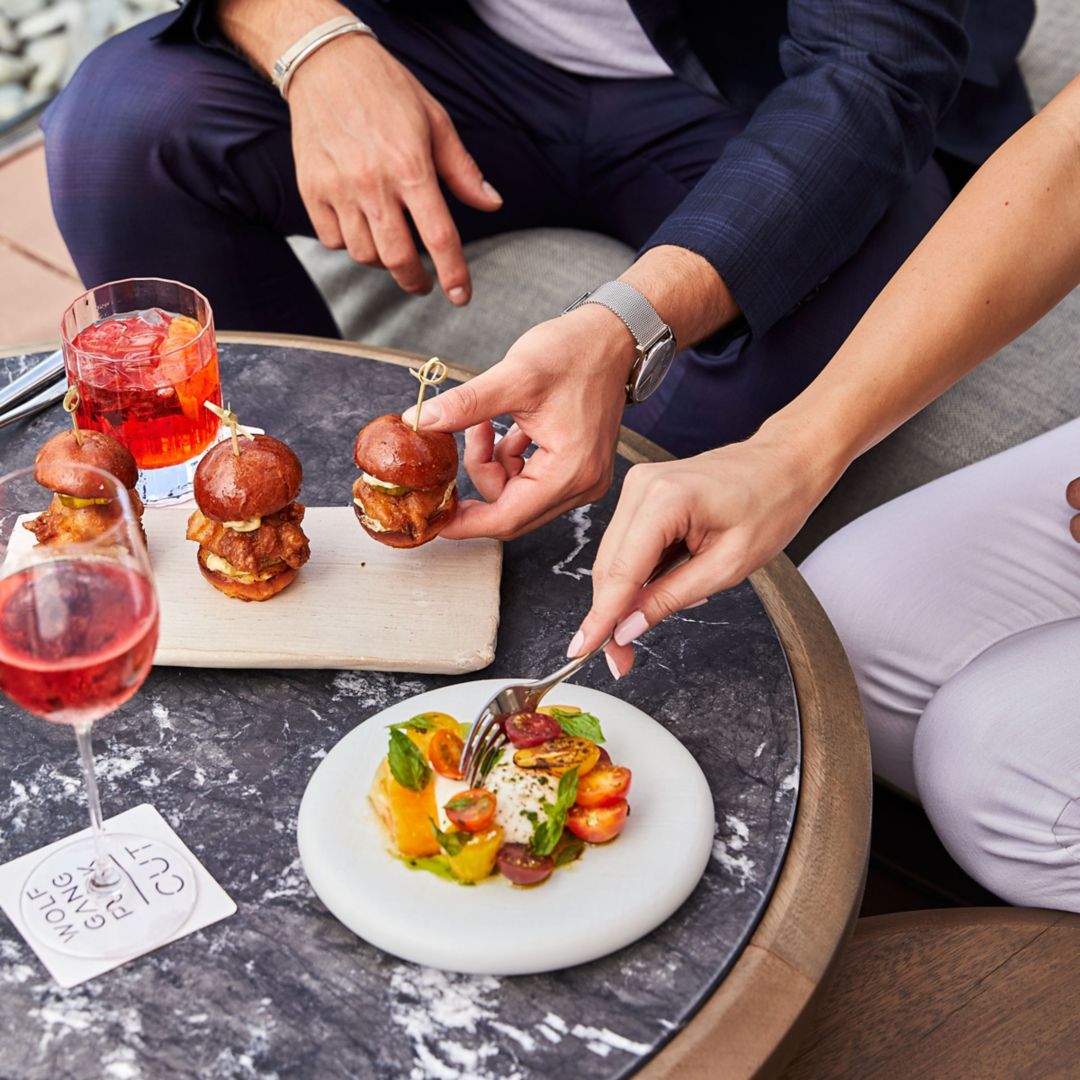 Two cocktails, a plate with sliders, and a plate of burrata with hands grabbing bites.